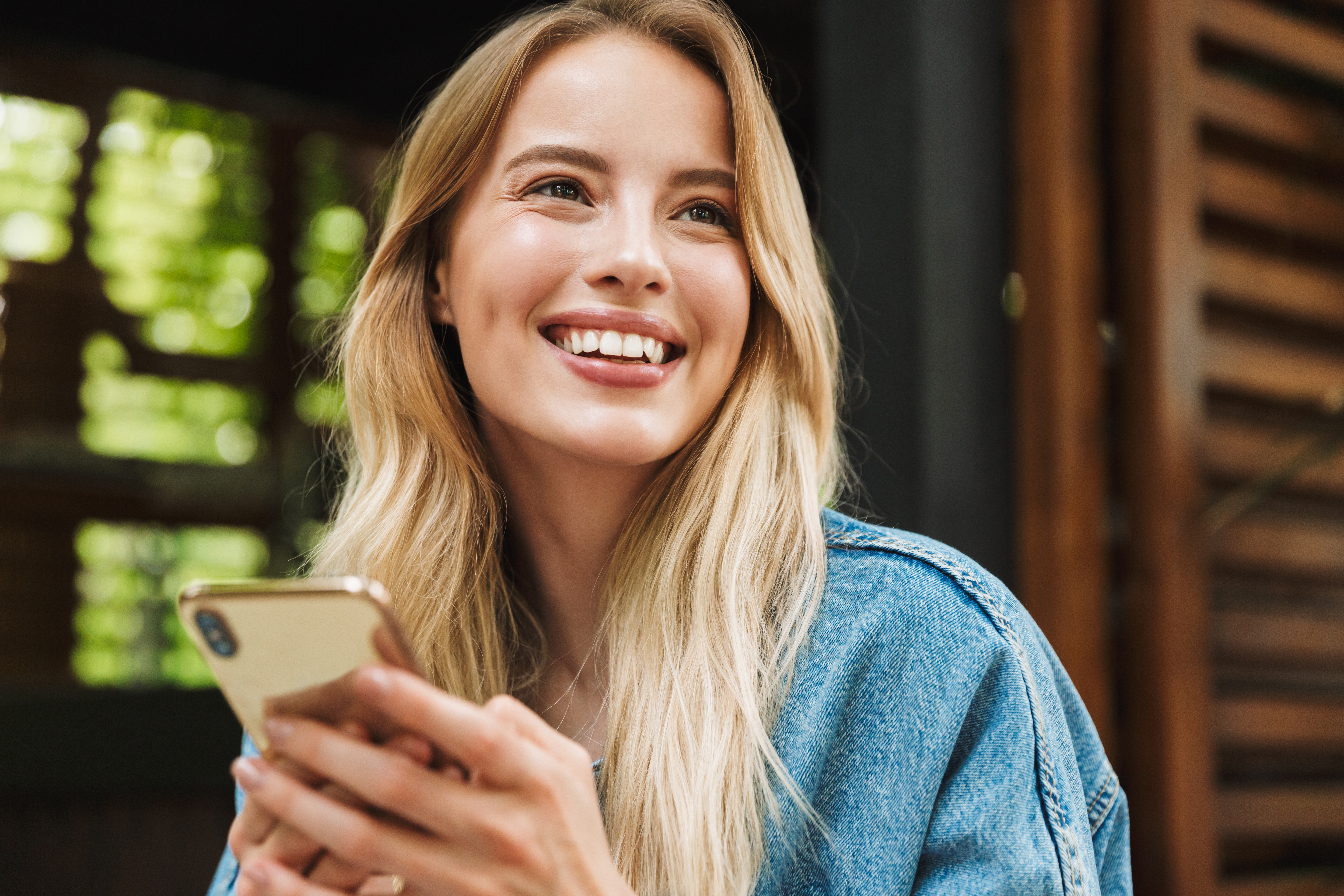 Junge Frau lächelt und hält ein Handy in der Hand, im Hintergrund ist eine Glasscheibe zu sehen.