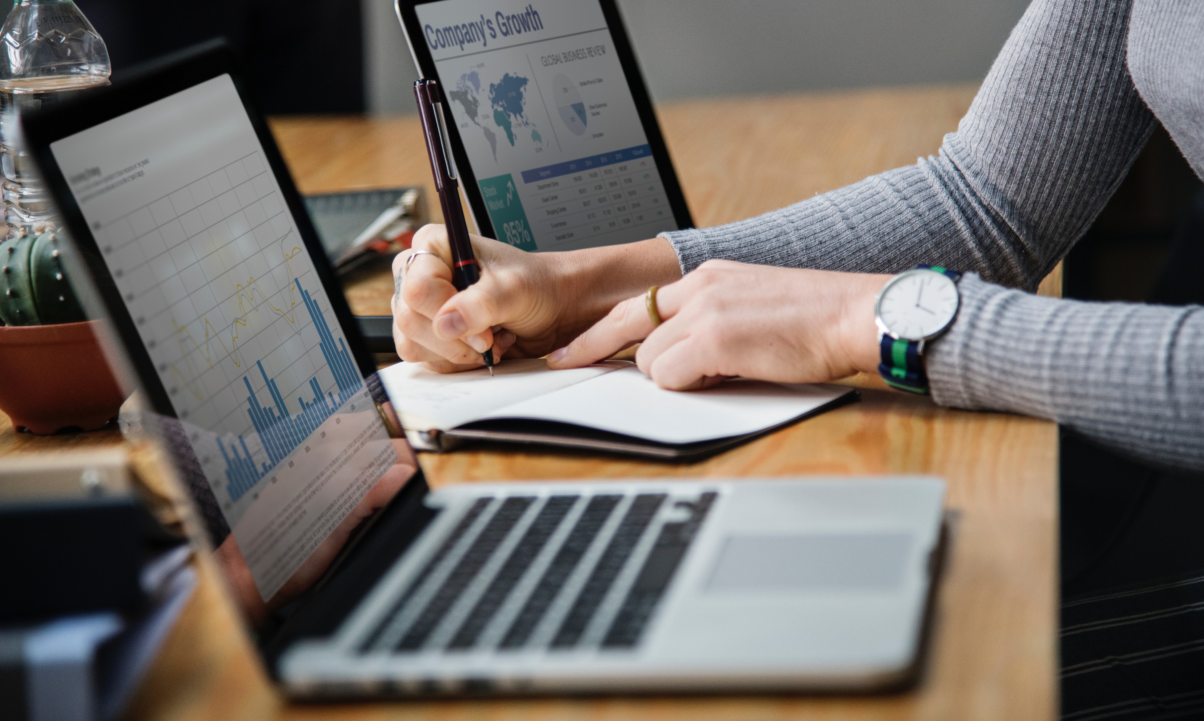 Zwischen zwei Laptops auf einem Tisch schreibt einer Person etwas auf einen Zettel.