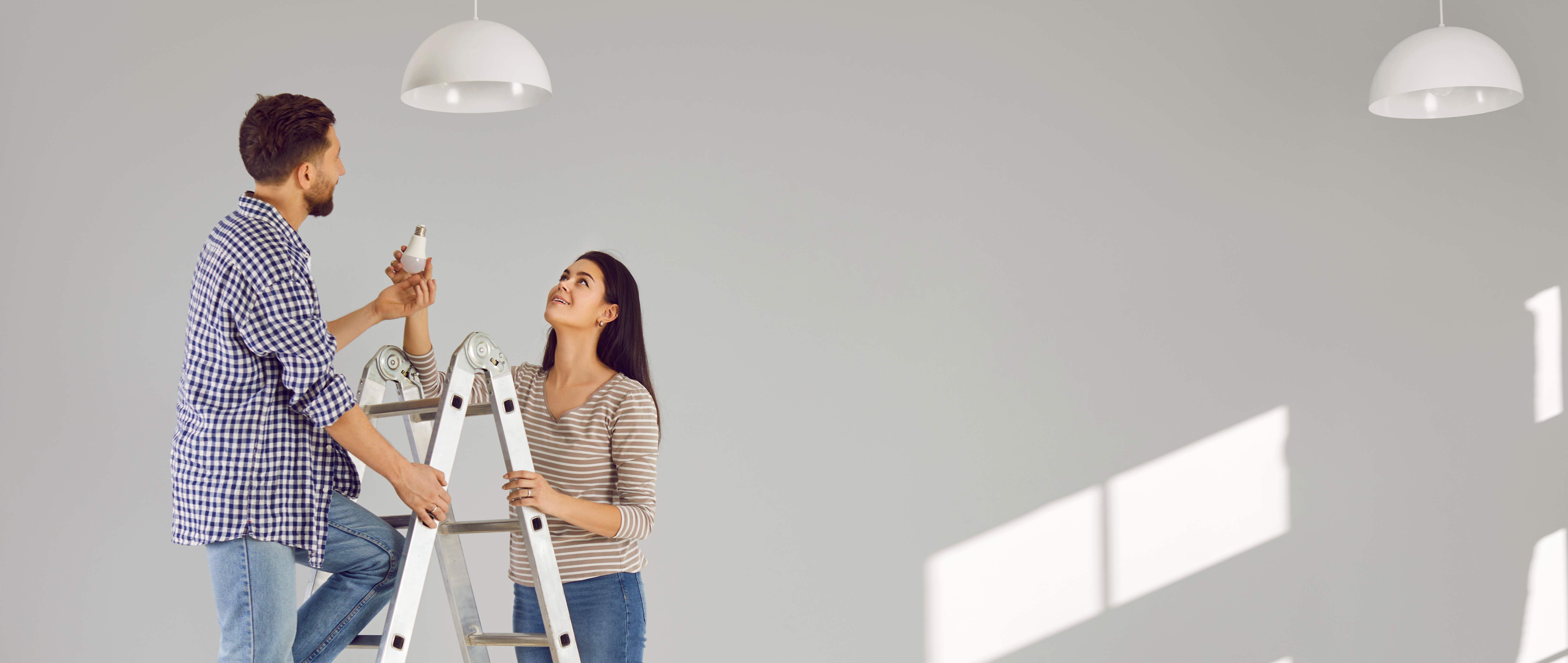 Ein Mann steht auf der Leiter in einer leeren Wohnung und möchte eine Glühbirne in eine Lampe drehen. Eine Frau steht daneben und hält die Leiter fest.