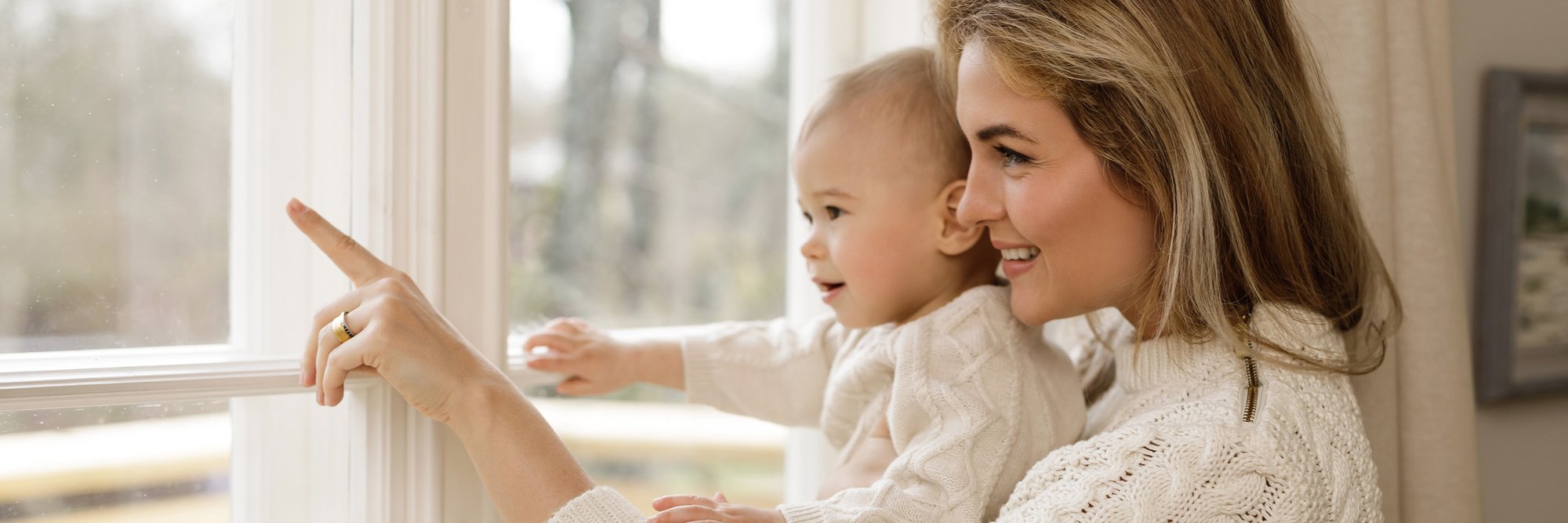 Mutter zeigt mit Baby auf etwas vor dem Fenster. Das Bild zeigt eine Mutter, die mit ihrem Baby vor einem Fenster steht und auf etwas zeigt. Beide wirken neugierig und entspannt.