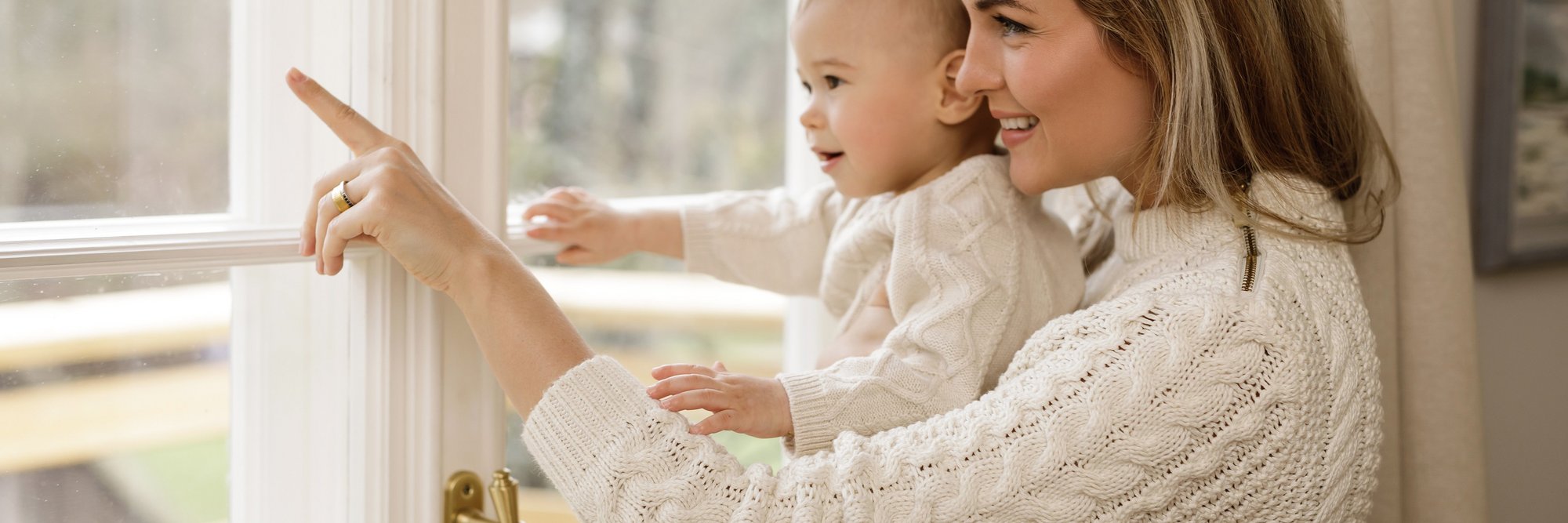 Mutter zeigt mit Baby auf etwas vor dem Fenster. Das Bild zeigt eine Mutter, die mit ihrem Baby vor einem Fenster steht und auf etwas zeigt. Beide wirken neugierig und entspannt.