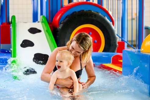 Frau und Kind im Wasserspielbereich. Eine Frau und ein Kind spielen fröhlich im Wasserpark, umgeben von spritzendem Wasser und bunten Rutschen.