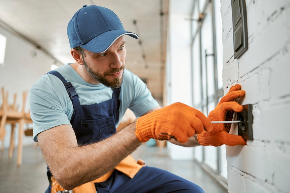 Handwerker installiert Steckdose an der Wand. Das Bild zeigt einen Handwerker, der in Arbeitskleidung und mit Handschuhen an einer Wand eine Steckdose installiert.