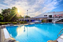 Das Bild zeigt ein Schwimmbad im Freien, das von der Sonne beleuchtet wird. Das Wasser glitzert im Sonnenlicht, und um das Becken herum gibt es grüne Bereiche und moderne Gebäude. 