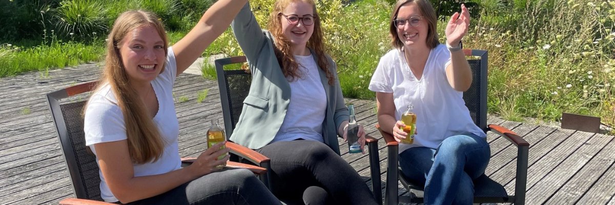 Das Bild zeigt drei Personen, die sich draußen auf einer Terrasse befinden und freundlich winken.