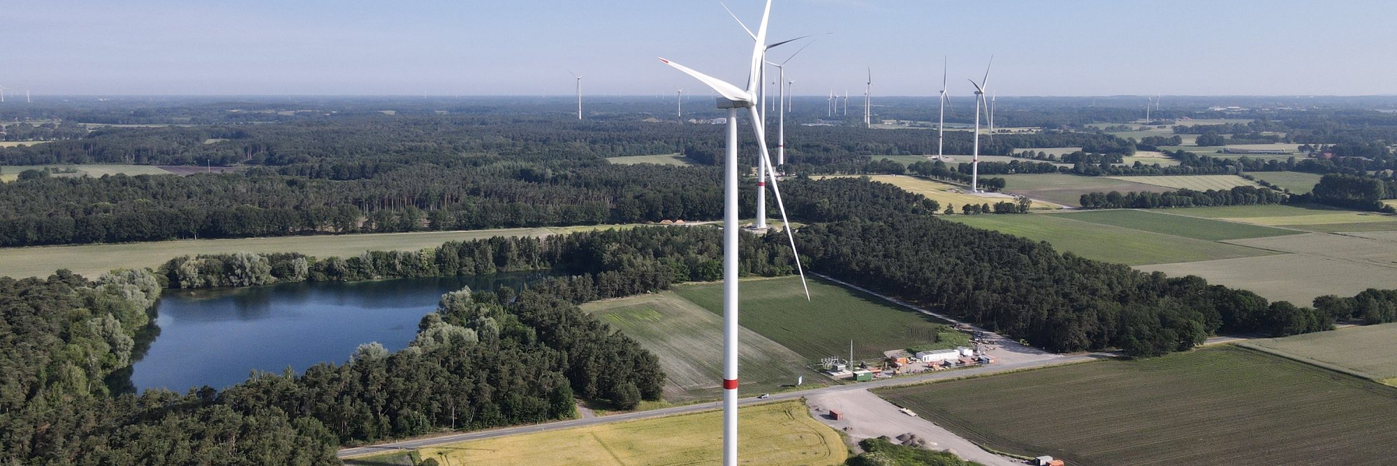 Luftaufnahme von Windkraftanlagen auf einem weiten, grünen Feld, umgeben von Bäumen und einem See.