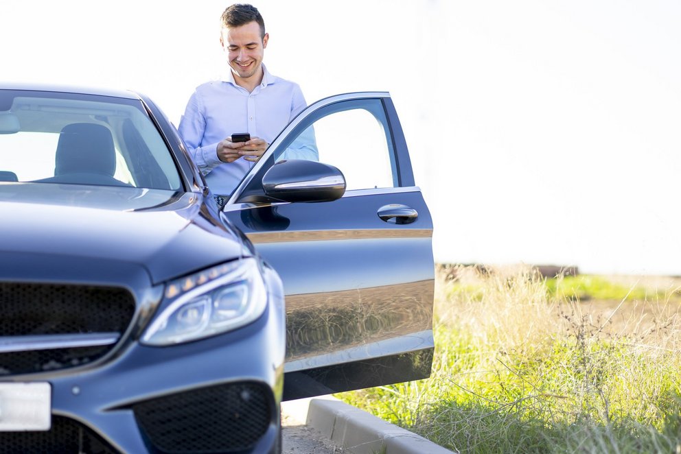 Person steht neben Auto und nutzt Smartphone. Eine Person steht neben einem Auto und nutzt ein Smartphone, während die Autotür geöffnet ist.