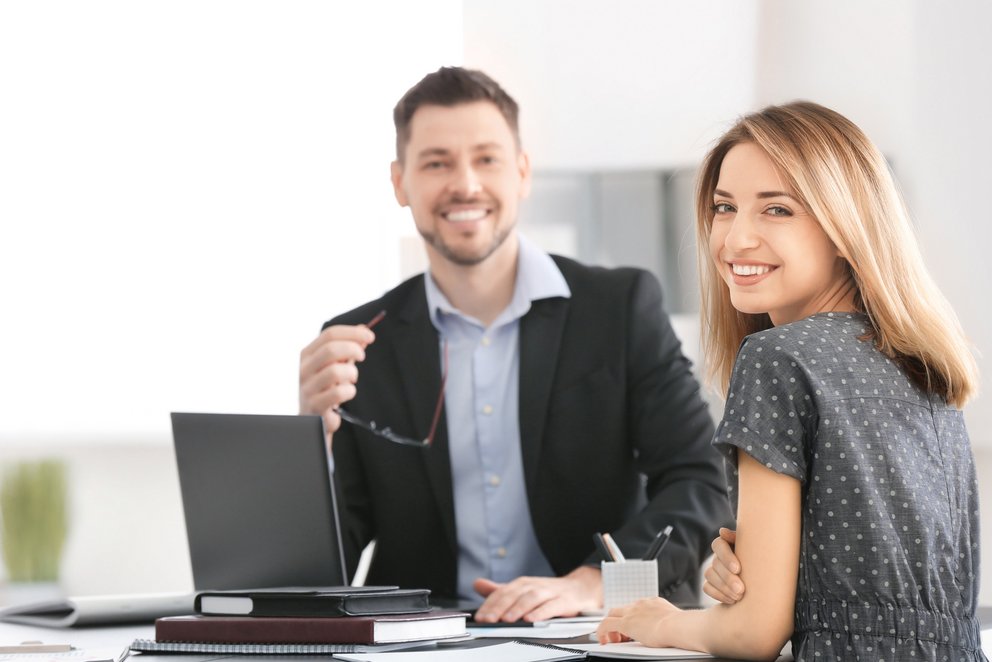 Freundliche Zusammenarbeit am Tisch. Die beiden Personen arbeiten zusammen an einem Tisch und schauen freundlich in die Kamera.