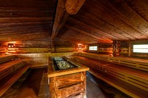 Das Bild zeigt das Innere einer traditionellen Sauna mit Holzverkleidung. Die Bänke sind in mehreren Ebenen angeordnet, und in der Mitte des Raums sind heiße Steine zu sehen.