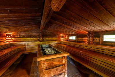 Das Bild zeigt das Innere einer traditionellen Sauna mit Holzverkleidung. Die Bänke sind in mehreren Ebenen angeordnet, und in der Mitte des Raums sind heiße Steine zu sehen.
