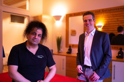 Das Bild zeigt zwei Personen in einem Wellnessbereich. Eine Person trägt ein schwarzes T-Shirt und steht vor einem Massagetisch, während die andere Person in einem Anzug daneben steht.