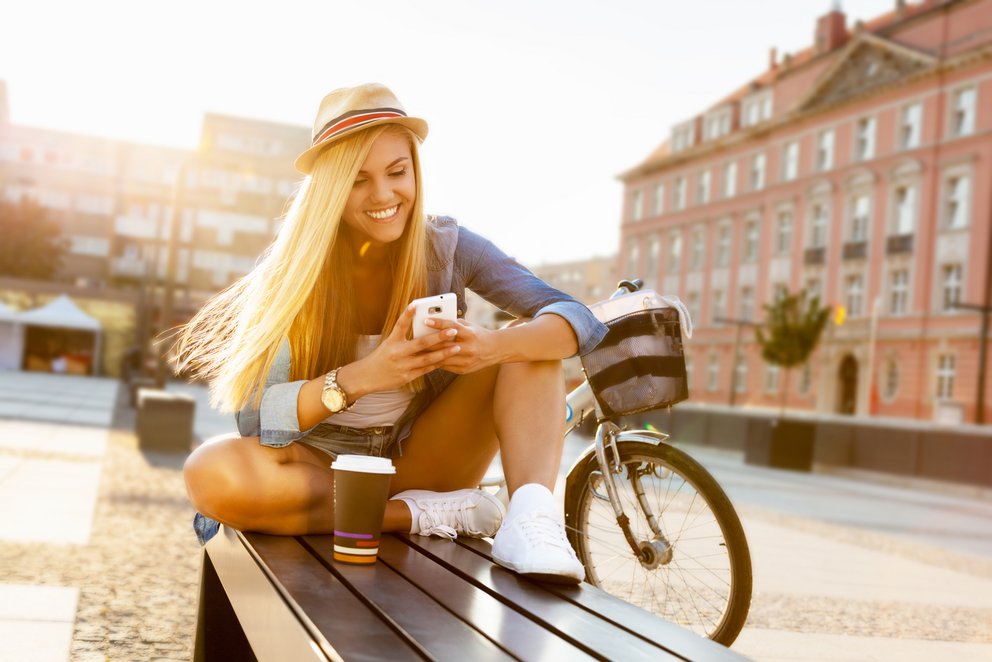 Frau auf Bank mit Handy und Fahrrad. Frau sitzt auf einer Bank und schaut auf ihr Handy, im Hintergrund ein Fahrrad.
