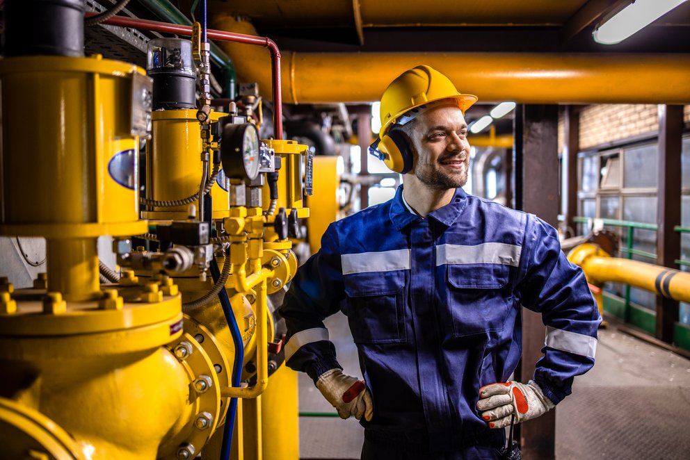 Techniker in Arbeitskleidung und gelben Helm steht vor Gasleitungen in einer Halle
