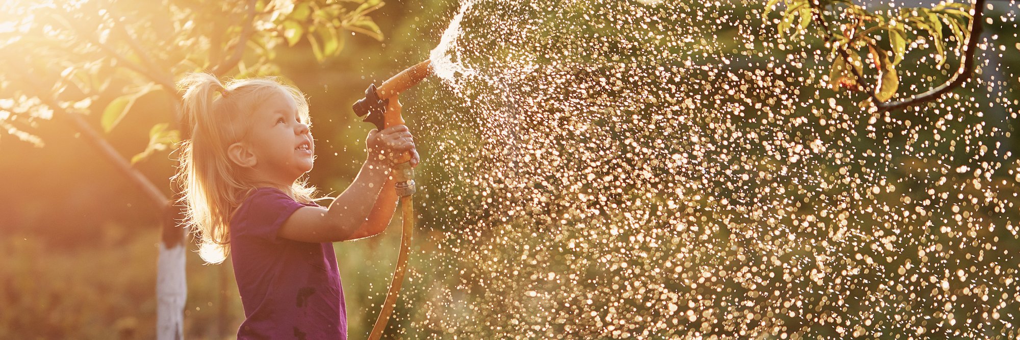 Kind spielt mit Gartenschlauch und spritzt Wasser. Das Bild zeigt ein Kind, das mit einem Gartenschlauch spielt und dabei Wasser in die Luft spritzt, das im Sonnenlicht glitzert.