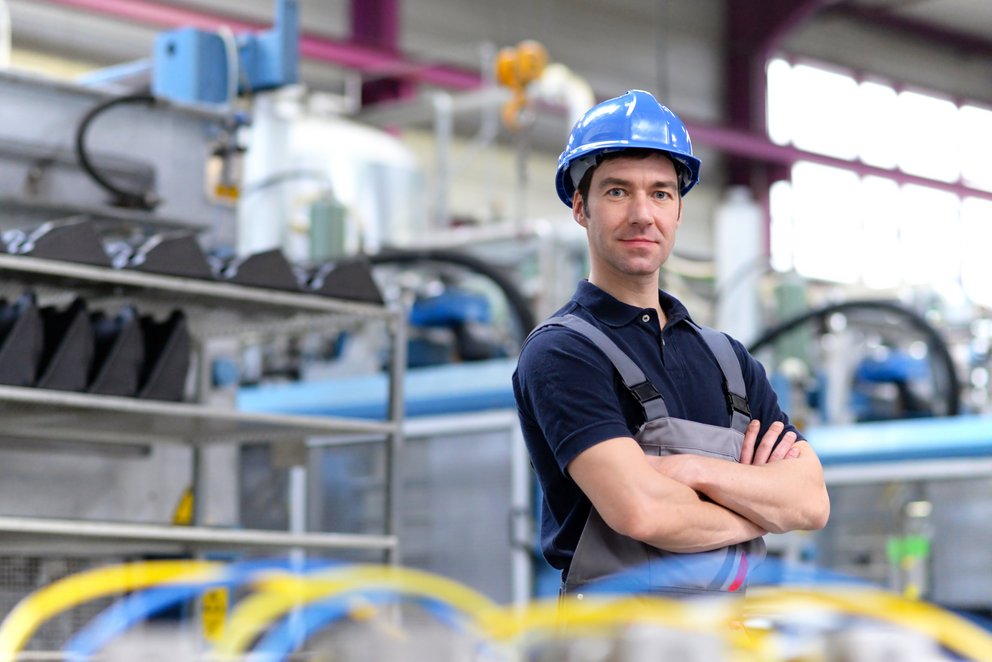 Person in Arbeitskleidung in der Fabrik. Das Bild zeigt einen Mann in Arbeitskleidung und Schutzhelm, der in einer Fabrik steht und die Arme verschränkt.