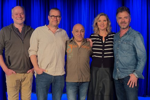 Fünf Personen stehen nebeneinander vor einem blauen Bühnenvorhang und lächeln in die Kamera.